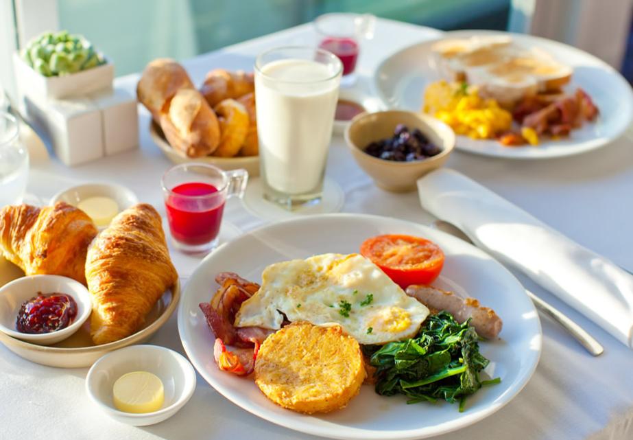 Colazione ricca: uova, croissant, succhi, latte. Atmosfera luminosa e invitante.