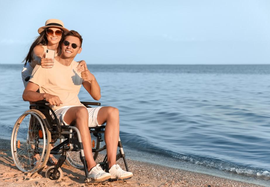 Coppia felice in spiaggia, lui in sedia a rotelle, sorrisi e abbracci al tramonto.